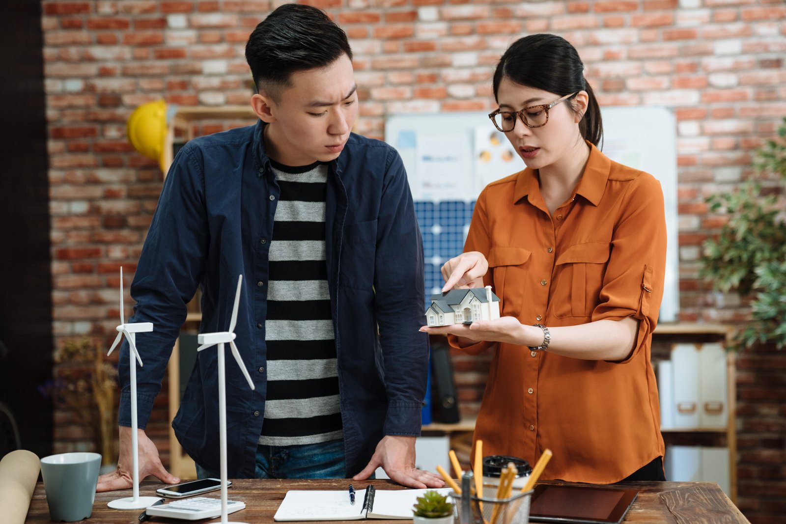 Group of young asian chinese architects with model of 3d house miniature standing in office and talking. frowning male coworker listening concentrated to female explaining on eco architecture.